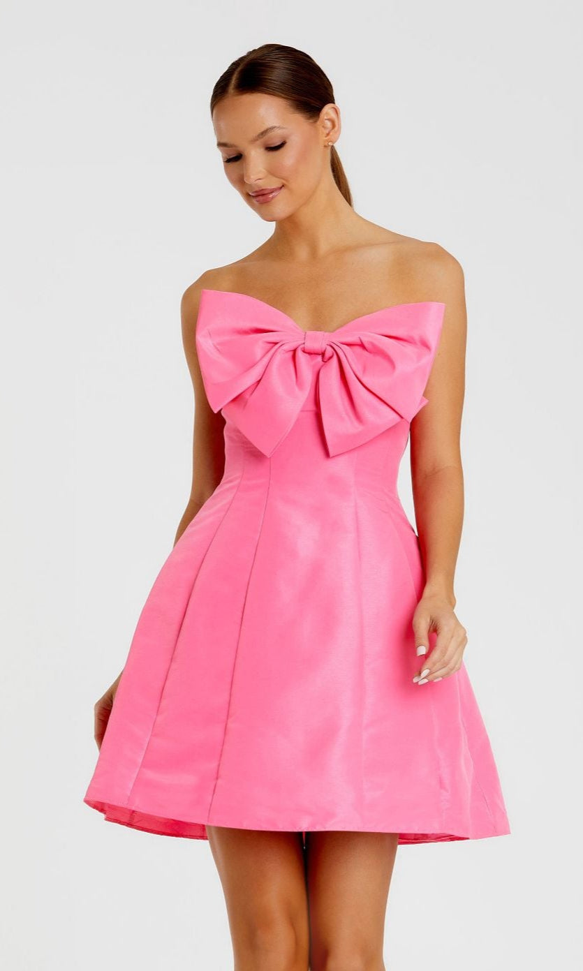 A woman models the Mac Duggal 49210 Strapless Short Bow Party Dress in bright pink, showcasing a bold chest bow, against a simple light background.